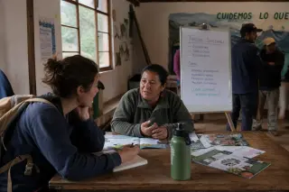 Travelers and local residents in conversation in a rural community hall, soft evening light, sustainable tourism discussion atmosphere